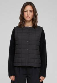Gilet matelassé noir avec boutons-pression dans un design boxy, doté de manches texturées. Le matériau semble léger et chaud, avec un look décontracté.