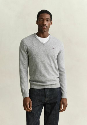 Uomo che indossa un maglione grigio chiaro a scollo a V sopra una T-shirt bianca e jeans blu scuro, in piedi davanti a uno sfondo beige uniforme.