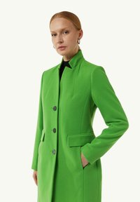 Grüner Wollmischungsmantel mit hohem Kragen, drei schwarzen Knöpfen und zwei seitlichen Taschen, der eine taillierte Passform und glatte Textur zeigt.