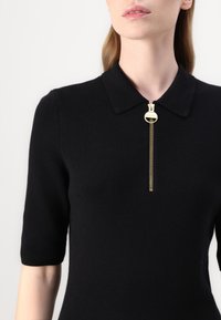 Haut en tricot noir à manches courtes, avec un col et un détail de fermeture éclair dorée. Texture lisse et silhouette ajustée.