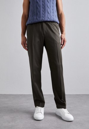 Osoba ubrana w ciemnooliwkowe spodnie o prostych nogawkach, białe trampki i niebieski bezrękawnik w warkoczowy wzór na jednolitym tle.