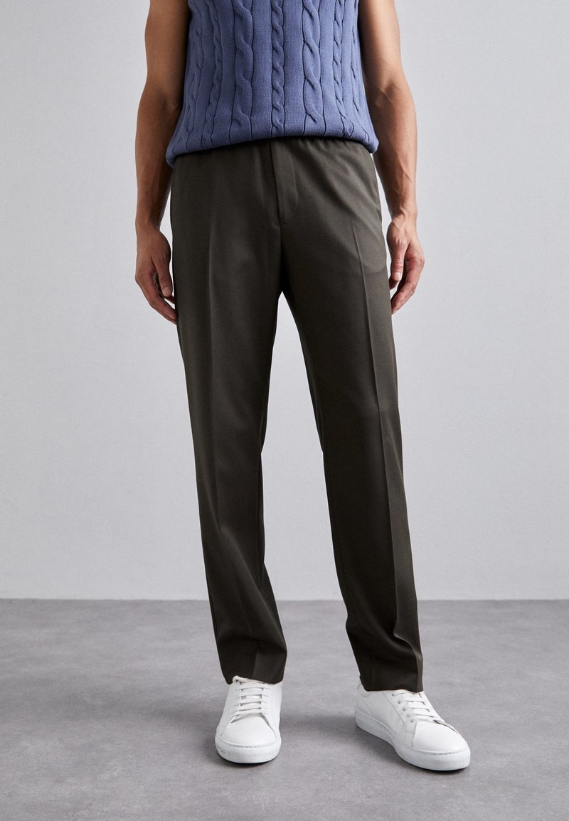 Personne portant un pantalon droit olive foncé, des baskets blanches et un pull sans manches en maille torsadée bleu sur un fond uni.