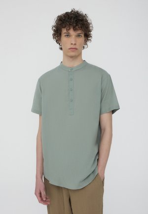Camicetta verde chiaro a maniche corte con colletto a banda, caratterizzata da una patta con bottoni frontali e una texture liscia, abbinata a pantaloni beige.