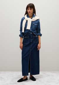 Camicia in denim con due tasche frontali, abbinata a una lunga gonna in denim blu con una fessura frontale. Sciarpa bianca a trecce drappeggiata sulle spalle, ballerine nere.