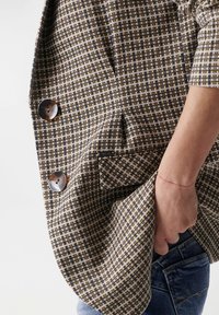 Blazer en mélange de laine avec un motif à carreaux marron, crème et marine, doté de deux boutons argentés et d'une poche latérale. Tissu texturé visible.