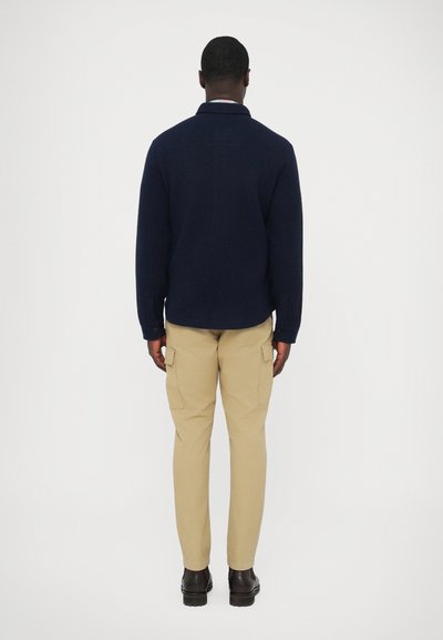 Camisola de malha azul-marinho com gola, combinada com calças cargo bege com bolsos laterais, usada com sapatos castanho-escuros.