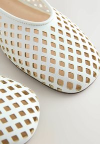 Ballerines blanches à enfiler avec des découpes en forme de losange sur la partie supérieure, laissant apparaître la doublure intérieure couleur beige clair et les bords de la semelle beige.