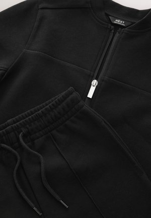 Schwarzes Sweatshirt- und Jogginghosen-Set. Das Sweatshirt verfügt über einen Reißverschluss und eine strukturierte Stoffoberfläche. Die Jogginghosen haben einen elastischen Bund mit Kordelzug.