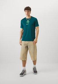 Teal short-sleeve t-shirt with a graphic logo and green accents, paired with light beige knee-length shorts and gray athletic shoes.