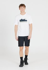 Valkoinen lyhythihainen t-paita, jossa on vuoristokuva ja teksti, yhdistettynä mustiin cargo-shorteihin, mustiin sukkiin ja tummiin vaelluskenkiin.