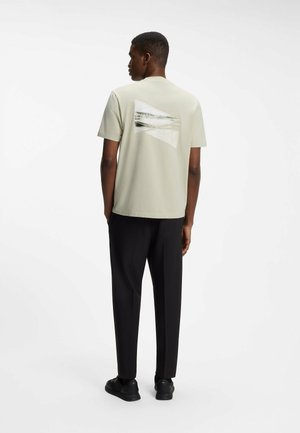 Mann steht mit dem Rücken zur Kamera, trägt ein beiges Grafik-T-Shirt, schwarze Hose und schwarze Sneakers vor weißem Hintergrund.