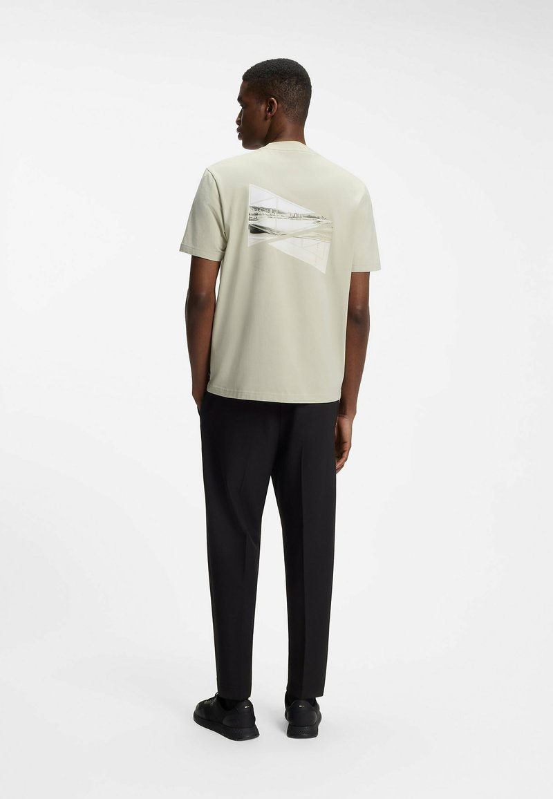 Homme debout, de dos, portant un t-shirt graphique beige, un pantalon noir et des baskets noires sur fond blanc.