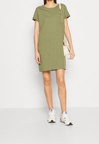 Robe courte olive à manches courtes avec un col rond et une poche poitrine. Portée avec des baskets blanches et beiges, présentant des motifs discrets.