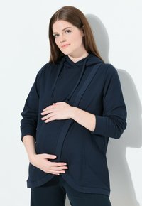 Donna incinta in un hoodie blu navy che culla delicatamente il suo pancione, in piedi contro un muro bianco, sorridendo dolcemente.