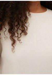 Prenda de tela blanca con una textura suave, que presenta un escote redondo y costuras visibles, complementada por cabello rizado y oscuro.