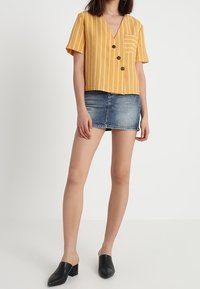 Gulrandig kortärmad topp med V-ringning och ficka, ihop med en denim minikjol och svarta klackskor. Knappar är framträdande detaljer.