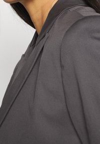 Ciemnoszara marynarka z gładkiej tkaniny, o wyraźnie zarysowanych klapach i dopasowanym kroju. Widoczne przeszycia dodają subtelnych detali.