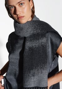 Sciarpa lavorata a maglia nera e grigia con un motivo ombre, presenta una texture morbida e le estremità frangiate, indossata sopra un top scuro con texture.