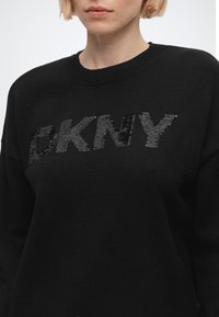 Gros plan d'une personne portant un pull noir avec un logo DKNY en sequins noirs brillants sur la poitrine.
