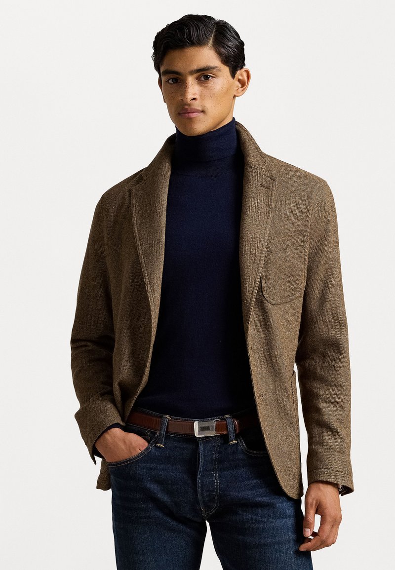 Brauner strukturierter Blazer mit Revers, getragen über einem marineblauen Rollkragenpullover, kombiniert mit blauen Jeans und einem braunen Gürtel mit einer silbernen Schnalle.