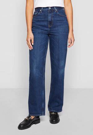 Kobieta ubrana w niebieskie jeansy o wysokim stanie i prostych nogawkach, biały top oraz czarne mokasyny z złotym łańcuszkiem, stojąca na szarej podłodze.