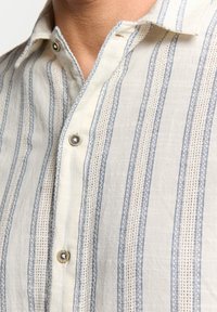 Lichtgewicht, crèmekleurig shirt met blauwe verticale strepen en subtiele textuurdetails. Voorzien van een button-down kraag en zilveren knopen.