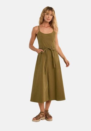 Donna che indossa un vestito midi verde oliva senza maniche con cintura in vita e sandali con plateau marroni con cinturini, in piedi davanti a uno sfondo semplice.