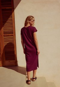 Robe violette longueur genou avec manches courtes et détail froncé au dos, associée à des sandales marron. Texture lisse contre un mur uni.