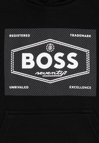 Tissu noir avec le texte blanc "BOSS seventy1" à l'intérieur d'un cadre hexagonal et les mots "Marque Déposée", "Excellence Inégalée" autour.