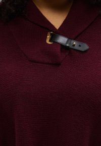 Bordový sveter s textúrovaným pleteným vzorom, ktorý má preložený golier a čierny kožený remienok so zlatou prackou ako dekoráciu.