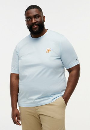 Uomo con barba che indossa una maglietta azzurro chiaro con un piccolo logo arancione sul petto e pantaloni beige, che posa con una mano in tasca su sfondo bianco.