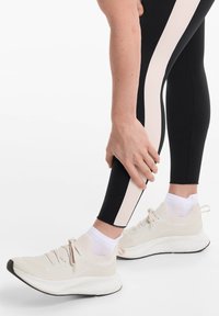 Fekete leggings bézs oldalsávval, bézs sportcipővel párosítva, melynek texturált felsőrésze és fehér talpa van. Fehér zoknik látszanak.