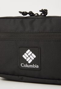 Juodasis "Columbia" maišelis su tekstūruotu audiniu, užtrauktuku ir išsikišusiu logotipo kuris pasižymi geometriniu dizainu.