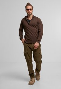 Hnědý pletený svetr s zipovým límcem, který je kombinován s texturovanými olivově zelenými cargo kalhotami. Outfit doplňují béžové boty s silnou podrážkou.