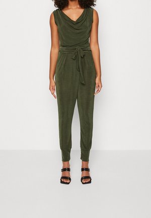 Kvinde iført en olivengrøn ærmeløs jumpsuit med draperet halsudskæring, knyttet talje, tilspidsede ben og sorte åbne tå-hæle.