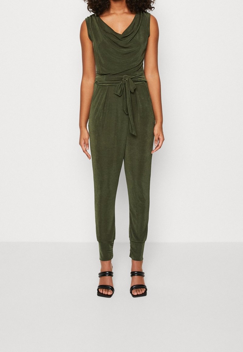 Femme portant une combinaison sans manches vert olive avec un col drapé, une taille nouée, des jambes fuselées et des talons ouverts noirs.