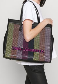 Bolso tote negro con un patrón de puntos multicolores, con franjas brillantes y el logo "KARL LAGERFELD" en negrita rosa en la parte frontal.