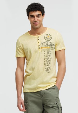 Mann mit lockigem Haar, der ein gelbes Henley-Kurzarmshirt mit grafischem Text und grüne Cargoshorts trägt, steht mit einer Hand in der Tasche.