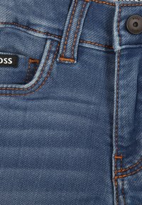 Modré džíny z denimu s oranžovým prošitím, vybavené páskem, knoflíkovým zapínáním a viditelnou visačkou s názvem značky.