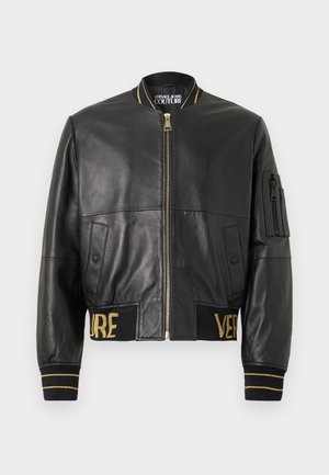 Must mustriga mustriga mustriga mustriga, millel on kuldsed dekoratsioonid, lukuga sulgemine, küljetaskud, soonikpihikud ja -vöökoht, millel on kuldne "VERSACE" broderi.