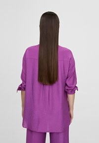Camisa de corte holgado y texturizada en un vibrante tono púrpura con mangas remangadas, combinada con la parte inferior a juego, vista desde la parte posterior.