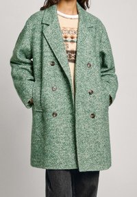Green herringbone coat with a double-breasted design, large lapels, and brown buttons. Soft texture, paired with a cream patterned sweater.