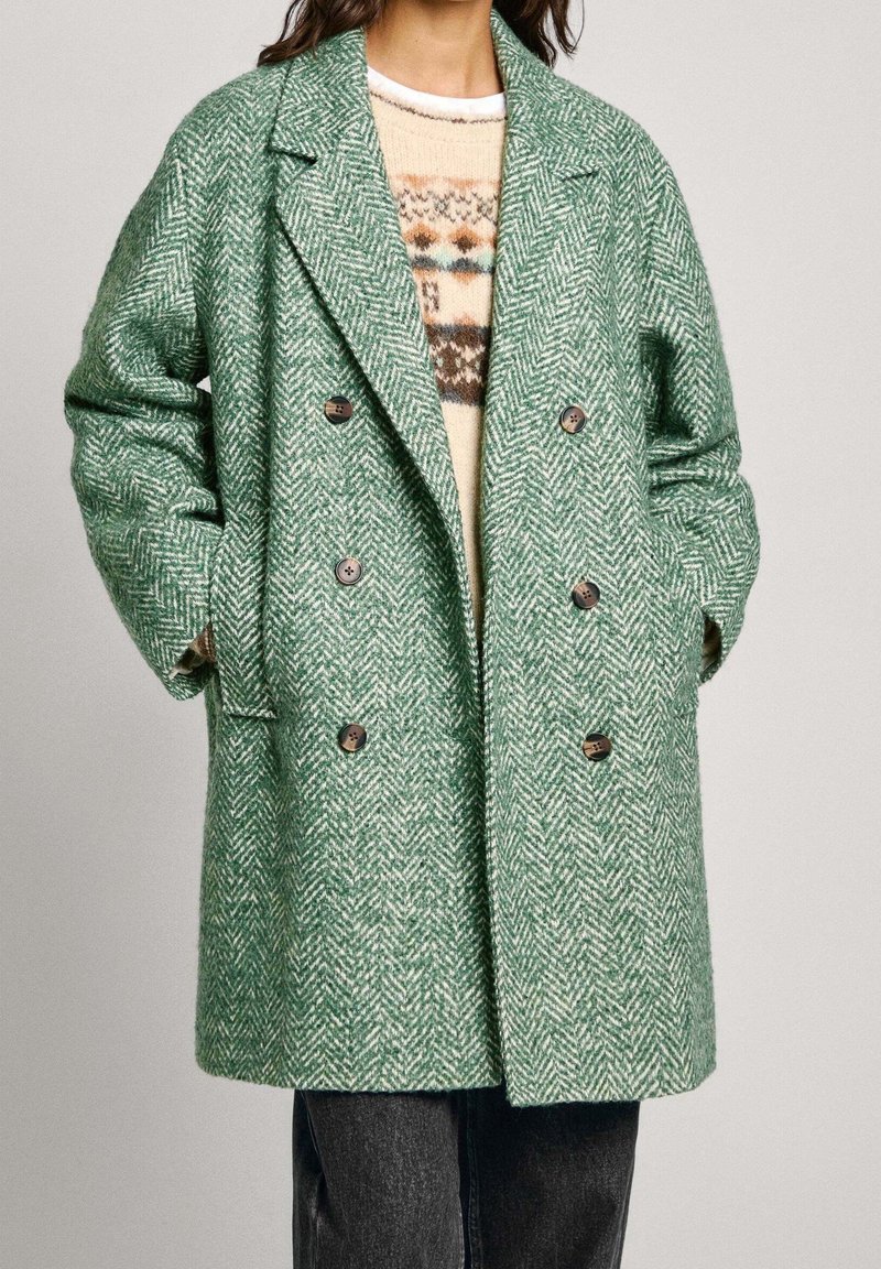 Green herringbone coat with a double-breasted design, large lapels, and brown buttons. Soft texture, paired with a cream patterned sweater.