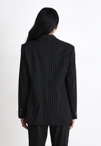 Blazer negru cu dungi, cu accente de fir metalic, umeri structurați, închidere cu un singur buton și manșete cu două butoane.