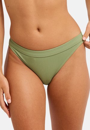 Femme portant un bas de bikini côtelé vert olive, tenant la ceinture latérale de la taille avec sa main droite.