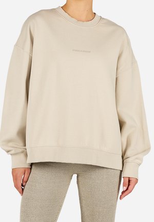 Frau trägt einen übergroßen beigen Sweatshirt mit Rundhalsausschnitt und gerippte beige Hose, steht vor einem einfarbigen Hintergrund.