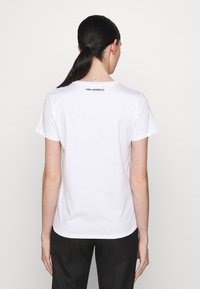 Biała bawełniana koszulka z okrągłym dekoltem i krótkimi rękawami, z nazwą marki "KARL LAGERFELD" nadrukowaną na czarno na górnej części pleców.