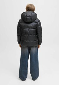 Doudoune noire avec capuche, présentant un design matelassé et une texture brillante ; associée à un jean large et décontracté bleu clair.