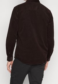 Camisa de pana marrón con mangas largas, puños abotonados y dobladillo curvado. Tejido texturizado con un acabado suave. Vista posterior mostrada.