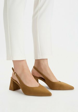 Stopy w brązowych zamszowych sandałach na słupku z odkrytym piętą i szpiczastymi noskami, zestawione z białymi dopasowanymi spodniami na jednolitym białym tle.